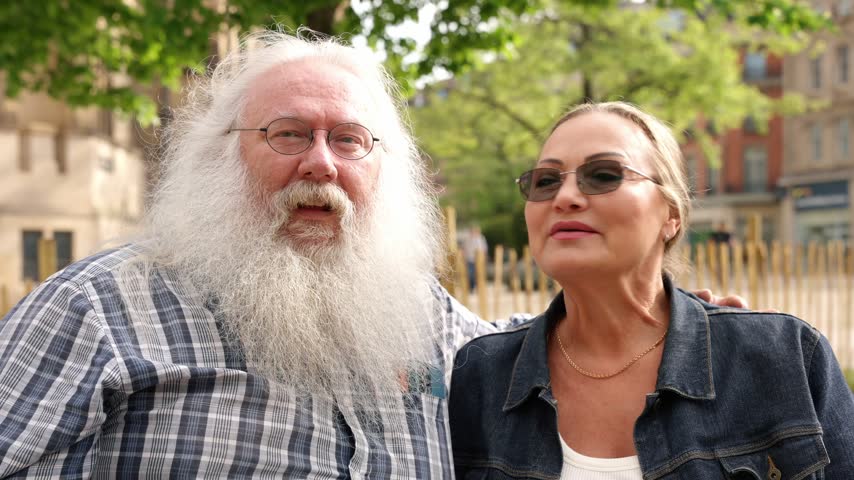 An older man with long white hair and a woman wearing sunglasses stand together on a tree-lined city street during the day.