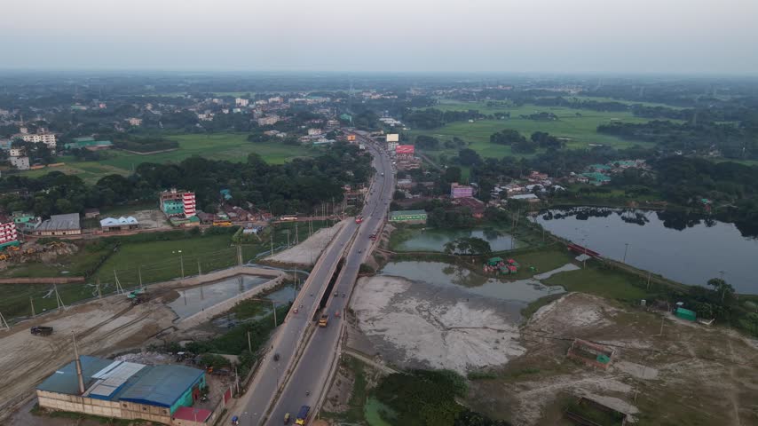 An elevated highway bridge stretches through a scenic landscape featuring ponds, lush green fields, and scattered residential buildings under a clear sky.