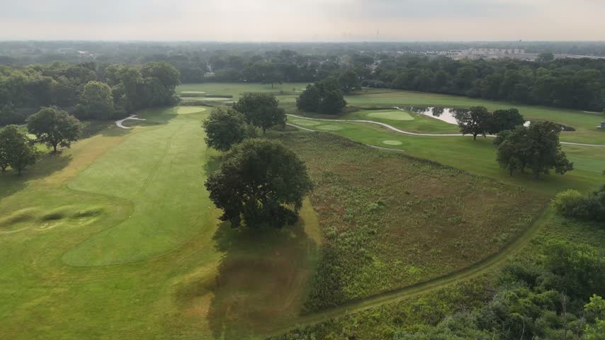 Wide aerial reveal over woods to a tree-lined golf course with bunkers under clouds. Filmed in River Grove, Illinois.
