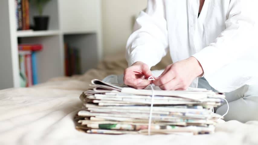 Person tying a stack of old waste newspapers with rope on a bed, preparing paper for recycling