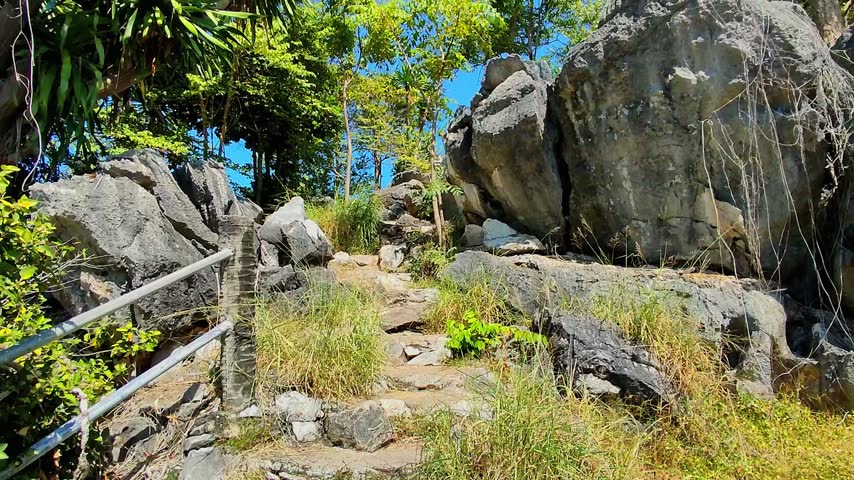 A stone path climbs between large rock formations and trees at an outdoor walking area during the day, with a metal handrail along the left side. Sam Roi Yot National Park in Thailand.