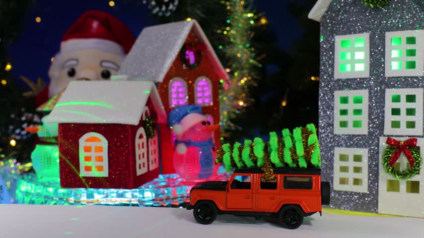 A yellow car pulls up to an orange car, some little houses spinning on a base, and a snowman holding a Christmas lantern