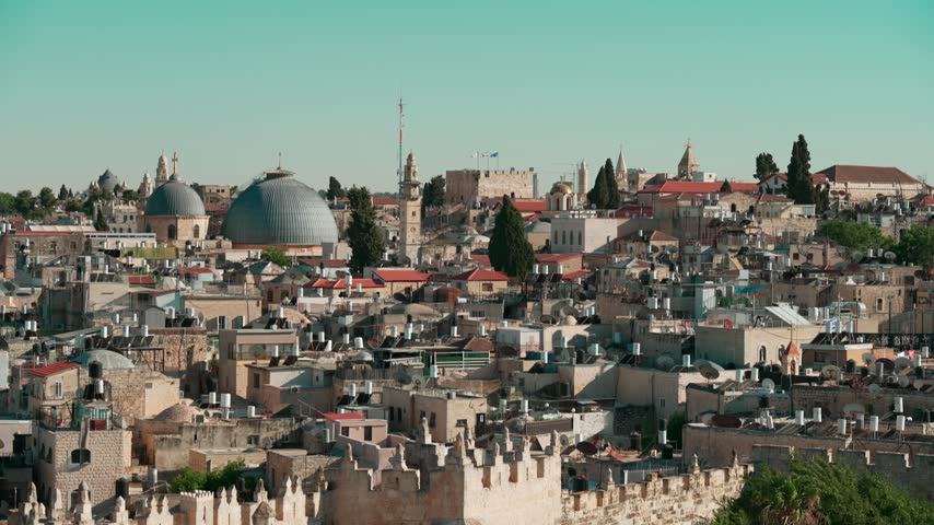 An afternoon telephoto view of East Jerusalem, where old Churches are intertwined with dense old houses carrying solar water heating systems, as filmed from a rooftop. few Israeli flags are seen.