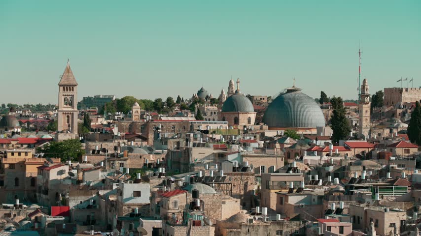 An afternoon telephoto view of East Jerusalem, where old Churches and Mosques are intertwined with dense old houses, as filmed from a rooftop. Israeli and Palestinian flags are seen at the distance.