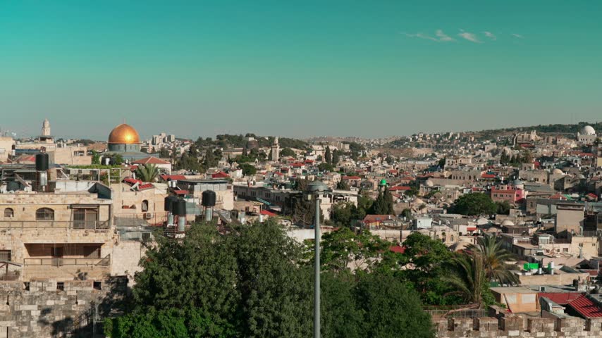 An afternoon view of East Jerusalem, where old Churches and Mosques are intertwined with old houses, as filmed from a rooftop. The Dome of the Rock is visible, golden shining and beautiful.
