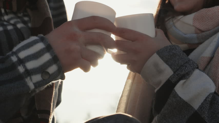 A young couple enjoys a hot beverage outdoors, dressed in striped and plaid scarves, celebrating a tender winter moment. Their smiles and gentle touch evoke warmth and connection