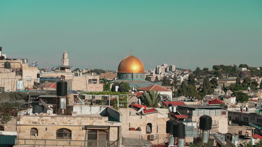The golden shining Dome of the Rock in an afternoon view of East Jerusalem, where old Churches and Mosques are intertwined with old houses. few Israeli flags are flapping in the wind.
