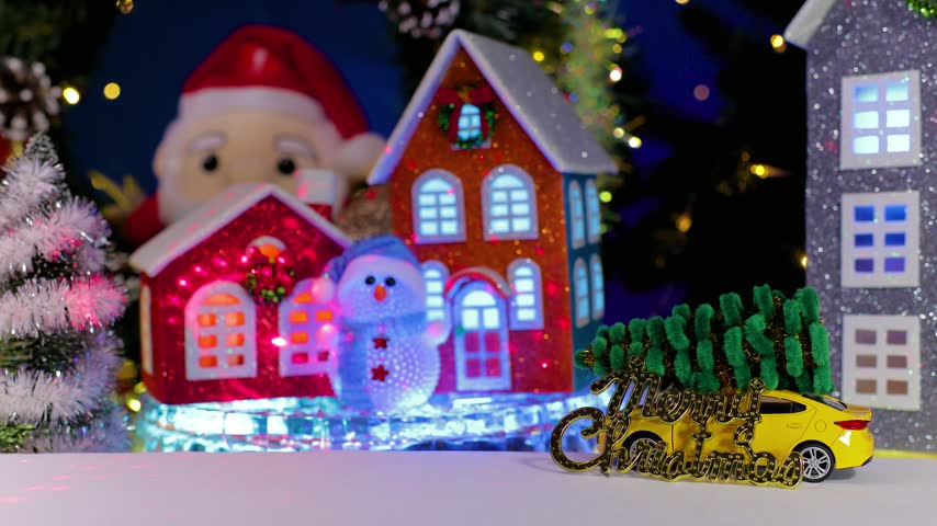 A yellow car pulls up to an orange car, some little houses spinning on a base, and a snowman holding a Christmas lantern