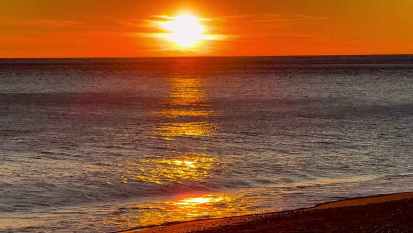 Bright orange sunset over the sea, with the sun near the horizon, gentle waves reaching the shore and golden sunlight reflecting on the water.
