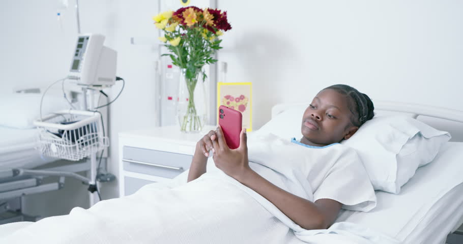 African American teen in bed wearing gown lifting red smartphone, reading, coughing, pressing chest. Hospital, patient, smartphone, medical, monitor, bedside, flowers