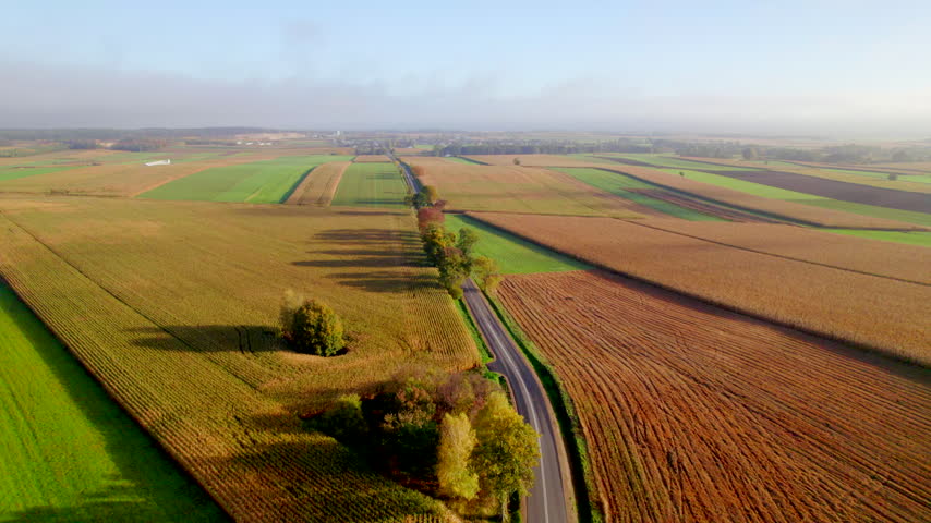 Aerial drone footage of a serene countryside landscape during autumn, featuring colorful fields, a winding road, and distant trees under a hazy sky