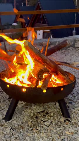 Close-up of bright orange flames burning wooden logs in a round metal fire pit outdoors, with A-frame cabins in the background.