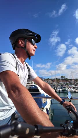 Mature man wearing a helmet and sunglasses while riding an electric bicycle alongside boats in the harbor, enjoying daily outdoor exercise and an active healthy lifestyle