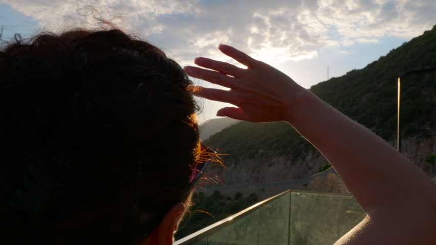 Woman watching the sunset over the mountains. Rear view of a young woman shielding her eyes with her hand while watching the beautiful sunset over the mountains, with bright sun rays