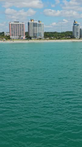 Vertical screen. Coastal view of Myrtle Beach, South Carolina, showcasing clear turquoise water and high-rise buildings along the shoreline