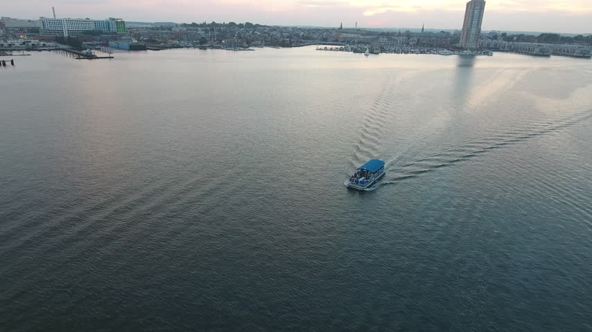 Aerial drone shot following a small water taxi boat leaving a long wake across calm Baltimore harbor water, then tilting up toward the waterfront skyline at dusk. Maryland, USA.