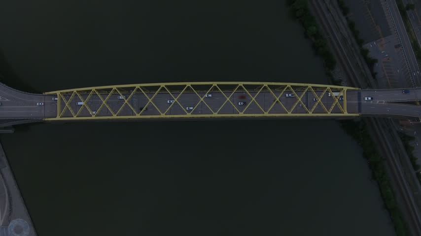 Top-down aerial drone shot hovering over a yellow steel bridge with cars crossing over the Allegheny River in Pittsburgh at dusk. Pennsylvania, USA.