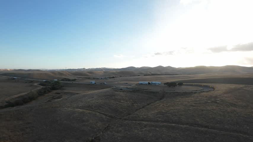 Scenic aerial view flying over the arid, rolling hills of the San Joaquin Valley in California, revealing a highway with traffic and distant mountains under a bright, setting sun