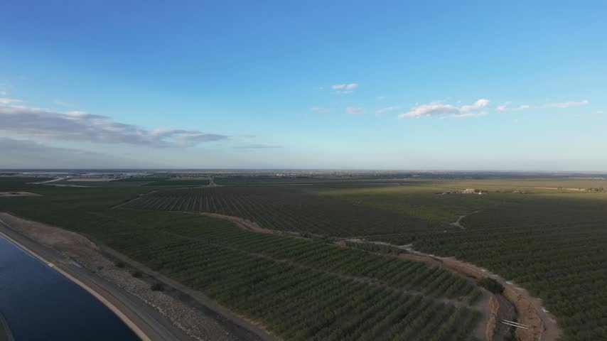 Panning aerial perspective following the California Aqueduct as it runs alongside lush agricultural fields and orchards in the San Joaquin Valley, highlighting vital water infrastructure