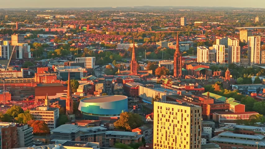 Coventry spires, the round blue building and tower blocks of the city centre glowing at golden hour in the West Midlands, England, aerial
