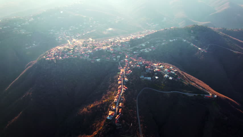 A mountain town spreads across a narrow ridge with dense red roofs and steep brown valleys on both sides while the drone approaches through hazy light revealing houses along the crest