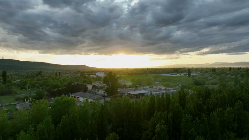 Aerial view of a small rural settlement in Georgia surrounded by green vegetation and open countryside. A dramatic sunset illuminates the horizon beneath thick clouds, while scattered homes