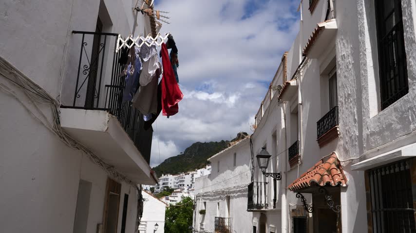 narrow alleyway in a traditional spanish white village with laundry hanging to dry on a balcony, white facades, wrought iron details, and mountain views under a blue sky in casares, andalusia, spain.