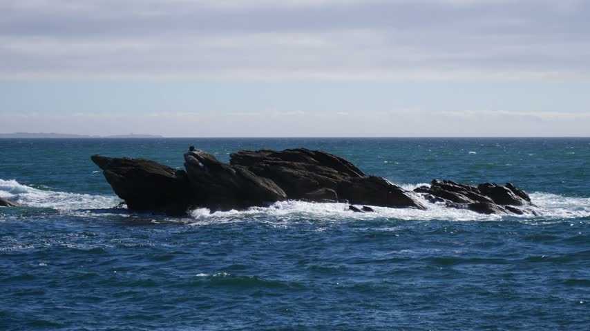  jagged dark rock formation sticking out of choppy blue ocean waters with white sea foam splashing against the coast under a overcast sky in Brittany France