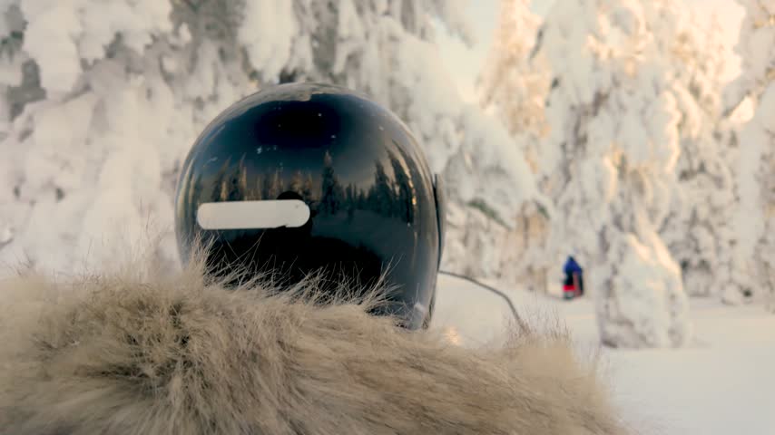 A glossy helmet rests on a snowmobile in a quiet forest of frost-covered trees in Lapland, Finland, during winter, with a rider visible in the distance.