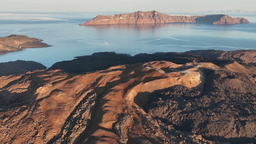 High aerial view shows volcanic terrain and geological formations of Nea Kameni in Greece with ocean and distant islands.
