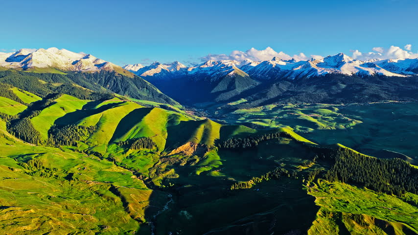 Panoramic high angle view of Kalajun grassland in Xinjiang with rolling green hills against snow-capped mountains.