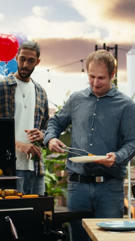 Vertical screen: Men talk and drink bottled beer beside an open rooftop barbecue grill where corn and patties are cooking, using utensils during a relaxed outdoor cookout and food preparation
