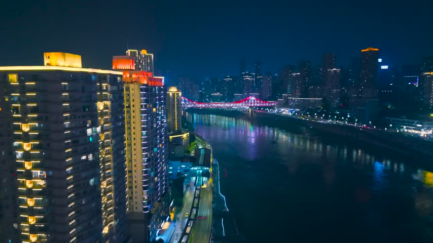 Aerial view of illuminated residential skyscrapers, traffic along the waterfront road, and a brightly lit bridge over the Jialing River at night in Chongqing, China.