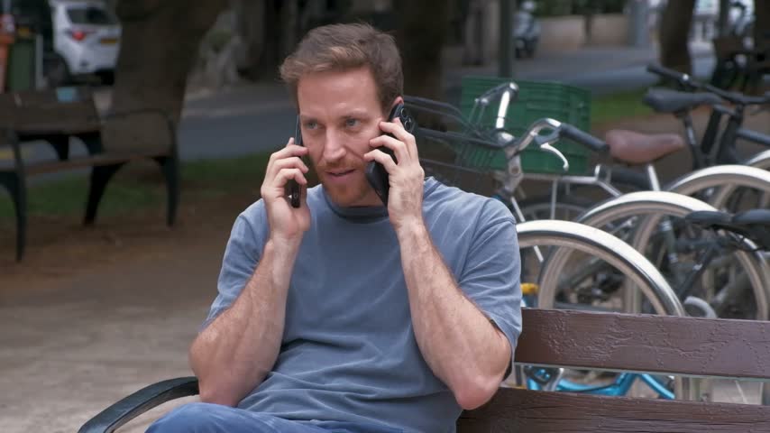 A man in a blue shirt sits on a wooden bench with a smartphone at each ear, talking beside parked bicycles in a playful illustration of multitasking and competing demands for attention