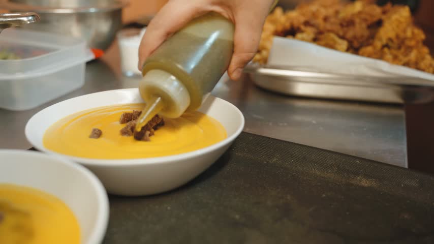 Squeeze bottle pouring a thin stream of olive oil over smooth orange vegetable soup topped with croutons in white bowls on a kitchen counter.