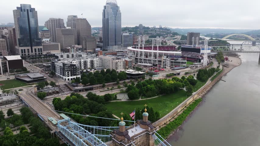 Roebling Bridge crosses the Ohio River below downtown Cincinnati and its riverfront stadium district, aerial descend reveal with stadiums in backdrop
