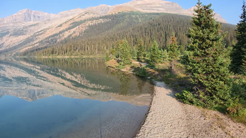 Bow Lake in Banff National Park reflects the Canadian Rocky Mountains and surrounding pine forests, creating a tranquil and symmetrical natural landscape under a clear sky in Banff NP, Alberta, Canada