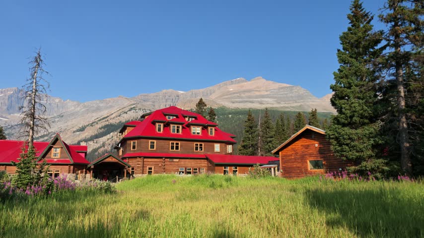 Num-Ti-Jah Lodge and adjacent cabins with vibrant red roofs sit in green meadow, surrounded by pine trees and the towering Rocky Mountains under clear blue sky in Banff National Park, Alberta, Canada.