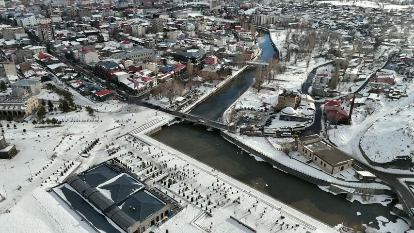 Aerial view of the snowy city of Kars with the Kars River, an old stone bridge and mountains on the horizon in winter, Turkey