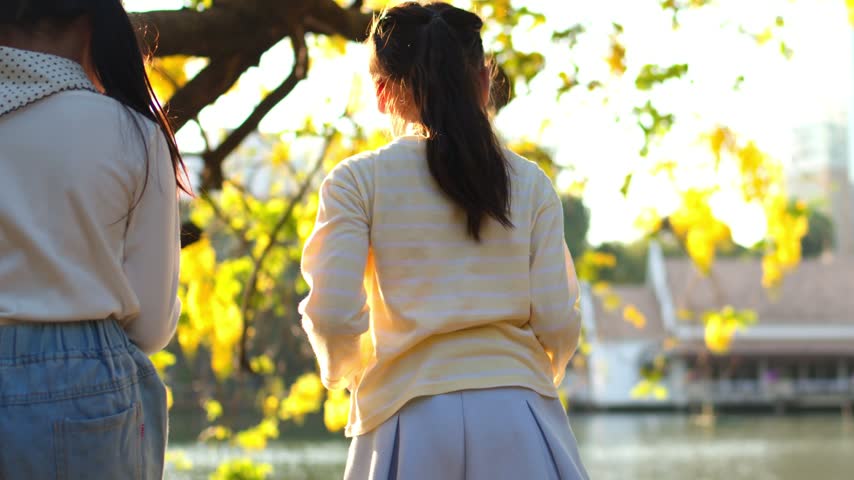 Slow zoom out of two schoolgirls chatting beside a sunlit lake in a city park.