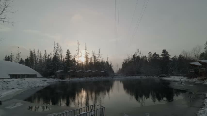 Sunrise Through Bare Trees Over Calm Lake And Snow-covered Cabins At Winter Resort In Heilongjiang, China. wide shot