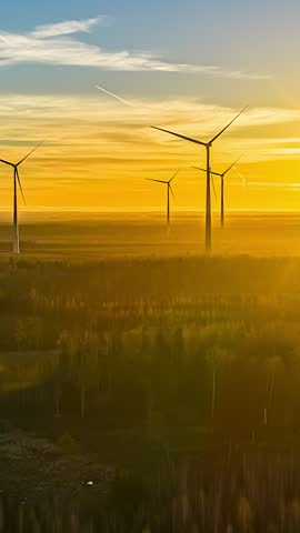 Wind turbines rise over a forested landscape beneath a golden sky streaked with clouds, vertical timelapse