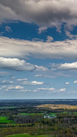 Clouds fill a blue sky over woodland, green fields and small ponds, vertical aerial time lapse