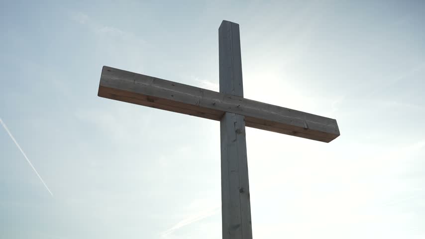 Bright sun shining from behind a wooden cross against a pale blue sky.