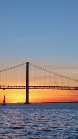 View of 25 de Abril Bridge famous tourist landmark of Lisbon connecting Lisboa and Almada over Tagus river with passing tourist yacht boat silhouette on sunset. Lisbon, Portugal.. Camera pan