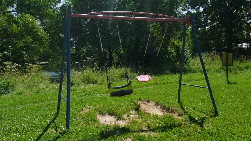 Swings are moving in a park under a bright sky with green grass and trees around