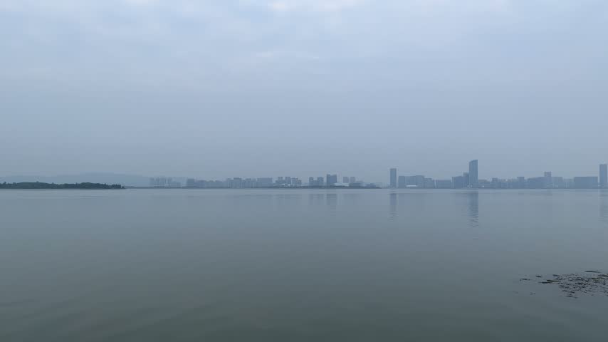 4K static wide shot of Taihu Lake and city skyline in Suzhou under overcast sky. Calm water reflects distant urban buildings and hazy atmosphere with gentle ripples.