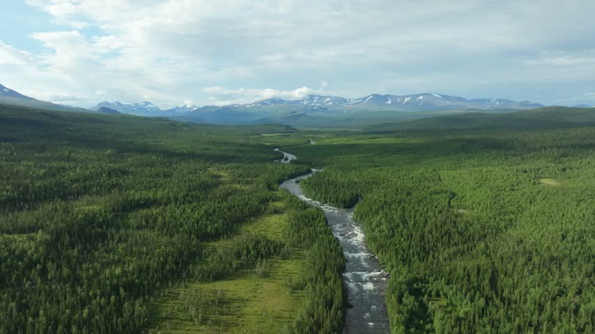 Aerial view of the winding Gamájåhkå river through a vast forest and valley, with snow-capped mountains in the distance along the Kungsleden trail in Sweden.