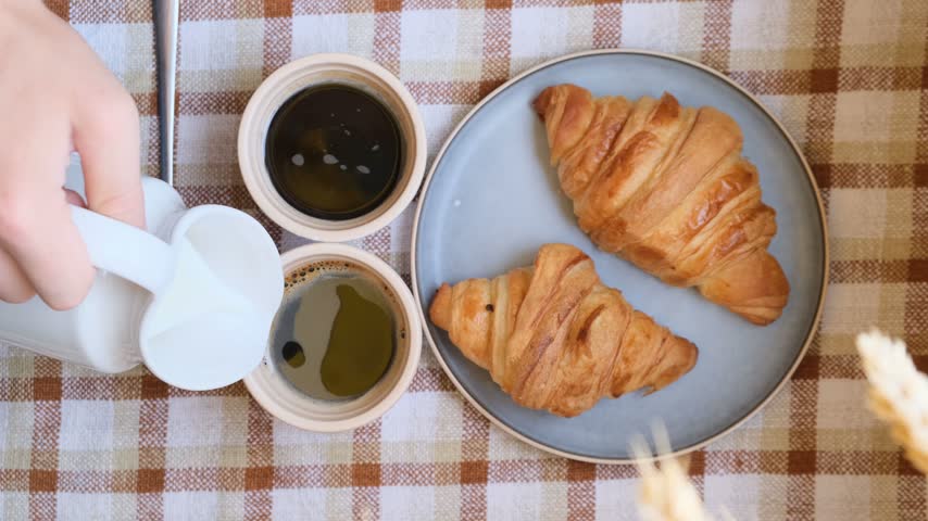 Top view of a hand using a ceramic pitcher to pour cream into two coffees beside fresh croissants, breakfast for two, oat milk alternatives, dairy products, workplace coffee break, fresh bakery.