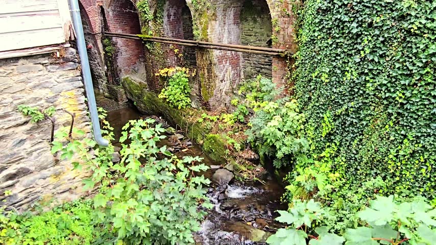 Small mountain stream flowing over stones next to historic brick arches covered with green ivy and wild vegetation. Picturesque rural architecture and natural scenery.